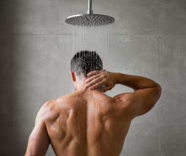 Athlete standing beneath a cold shower, shoulders tense, in a simple bright bathroom