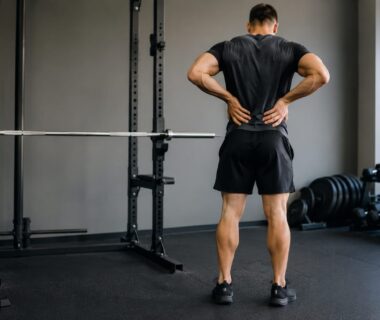 Strength athlete cautiously bracing lower back beside a squat rack during injury assessment