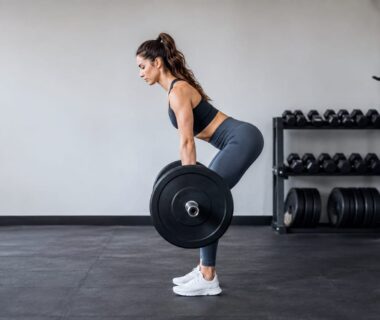 Side-view athlete demonstrating a controlled barbell hip hinge with neutral spine in a strength gym